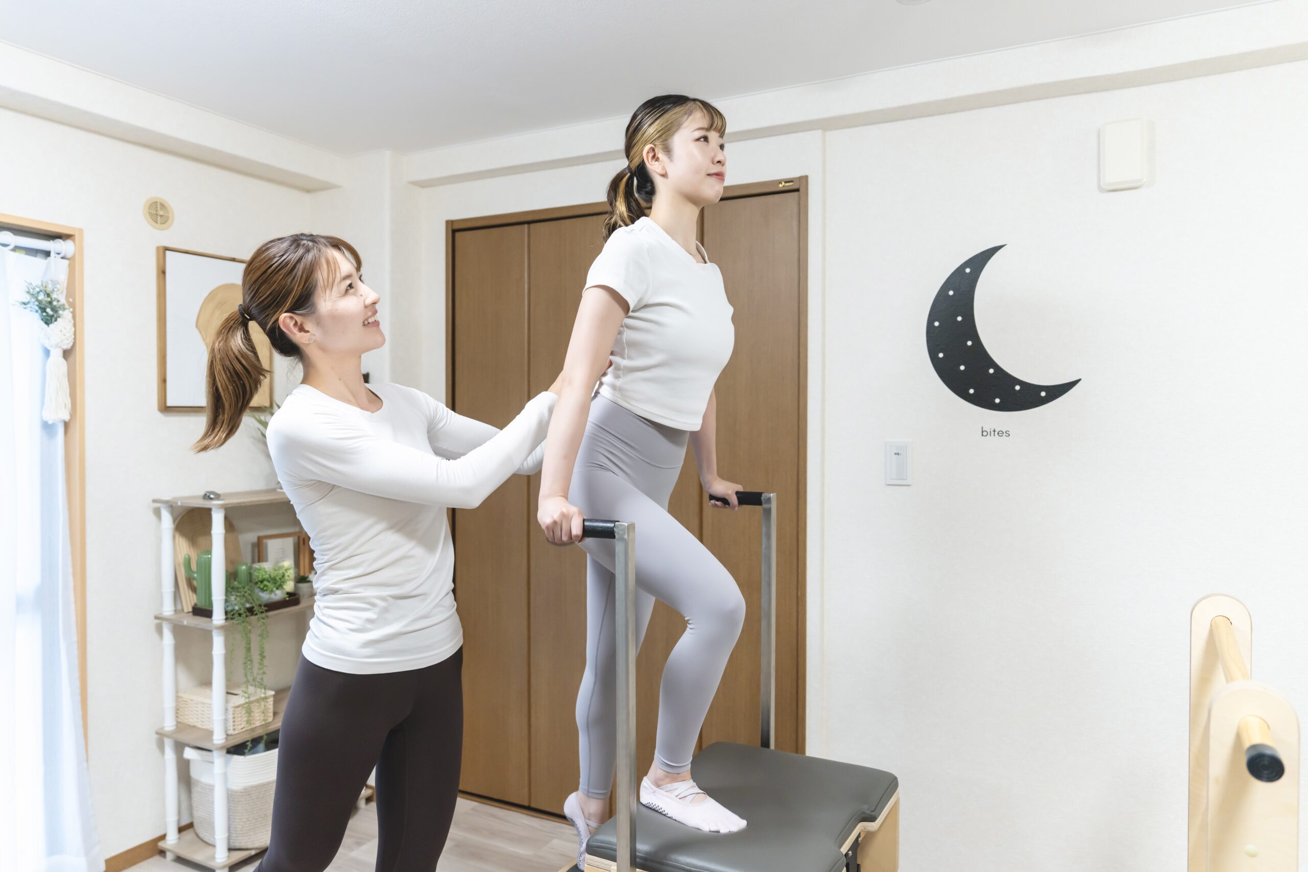 ピラティスマシンのバーを使って背筋を伸ばすポーズを行う女性と、それをサポートするインストラクター